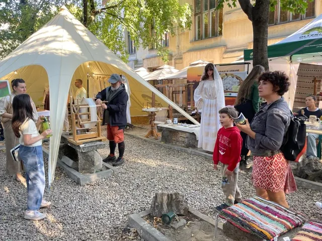 Oamenii stau în fața unui cort alb lângă un dispozitiv de țesut din lemn, în timp ce o femeie în rochie albă trece pe lângă ei.