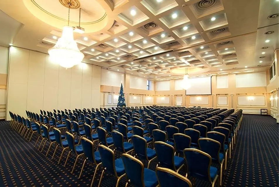 O sală de conferințe mare cu numeroase rânduri de scaune albastre dispuse pe un covor înfundat cu modele geometrice sub un tavan cu panouri luminoase.