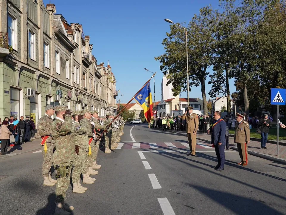 Soldați în uniforme de camuflaj stau în rând pe un drum asfaltat lângă un steag colorat, în timp ce civili și oficiali stau pe trotuar.