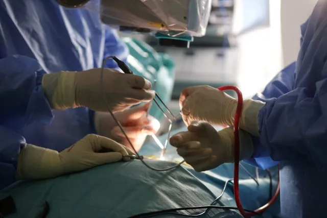 Mâini în mănuși sterile manipulează instrumente chirurgicale metalice deasupra unui pacient acoperit cu un drapou de culoare verde într-un cadru medical.