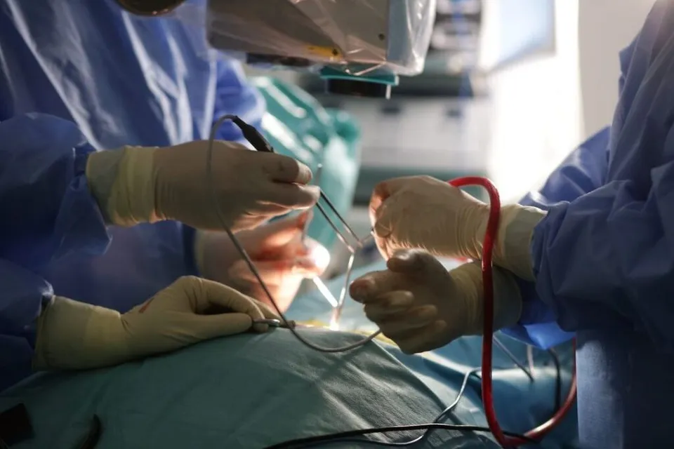 Mâini în mănuși sterile manipulează instrumente chirurgicale metalice deasupra unui pacient acoperit cu un drapou de culoare verde într-un cadru medical.