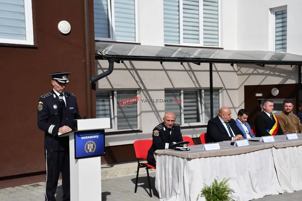 Un oficial în uniformă stă la un pupitru lângă un panou albastru, în timp ce patru bărbați stau la o masă acoperită cu un material alb.