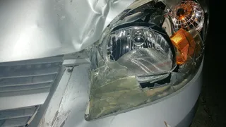 Un autoturism de culoare argintie prezintă o parte frontală deformată și un far stâng acoperit cu bandă adezivă transparentă.