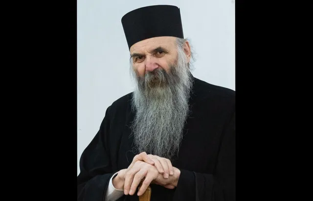 Un bărbat cu barbă lungă și gri poartă o pălărie neagră și o robă neagră, stând cu mâinile împreunate în față.
