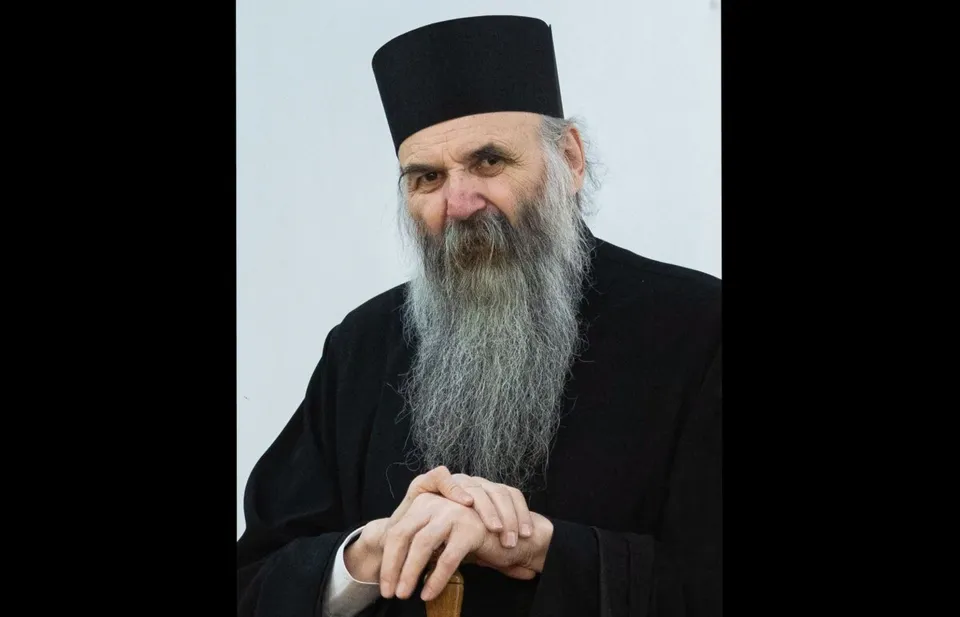 Un bărbat cu barbă lungă și gri poartă o pălărie neagră și o robă neagră, stând cu mâinile împreunate în față.