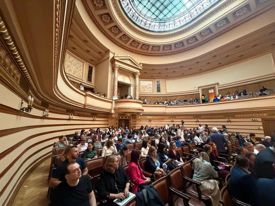 O mulțime de oameni stă așezată pe scaune în interiorul unui auditorium cu un dom circular și balcone decorate cu elemente clasice.