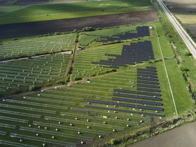 O vedere aeriană prezintă numeroase panouri solare dispuse în rânduri pe un teren verde lângă un drum.