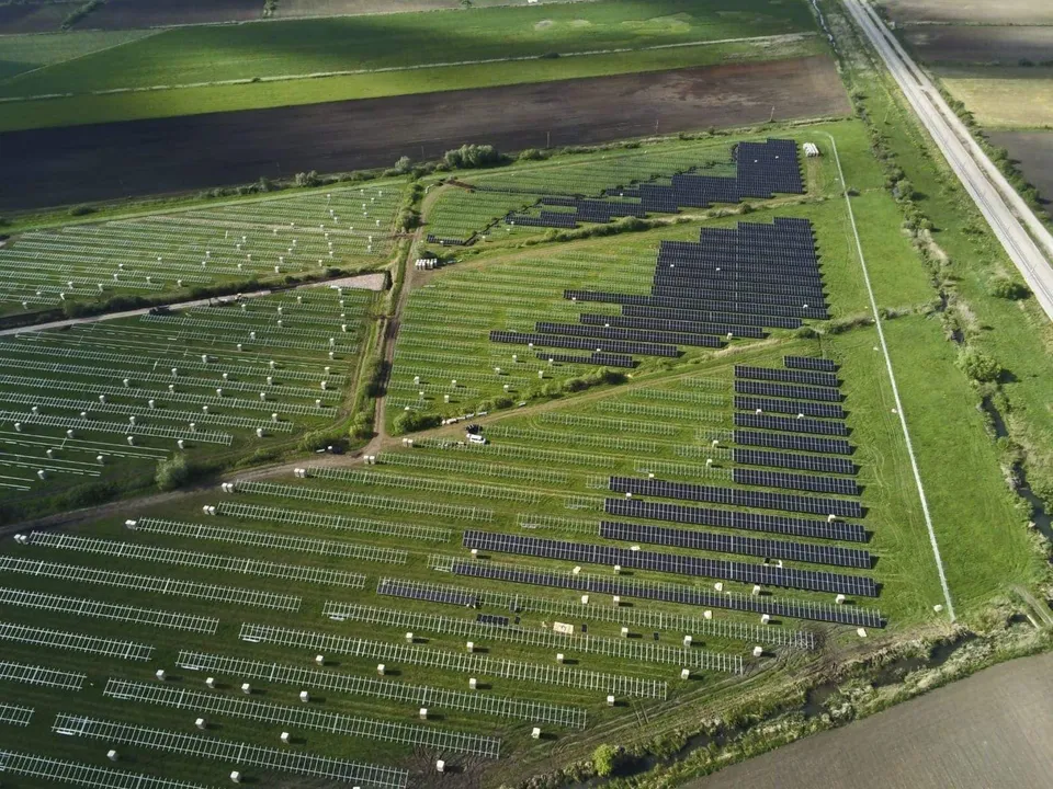 O vedere aeriană prezintă numeroase panouri solare dispuse în rânduri pe un teren verde lângă un drum.