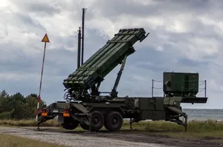 Un vehicul militar verde cu un lansator de rachete înclinat este poziționat pe un drum de pământ sub un cer înnorat.