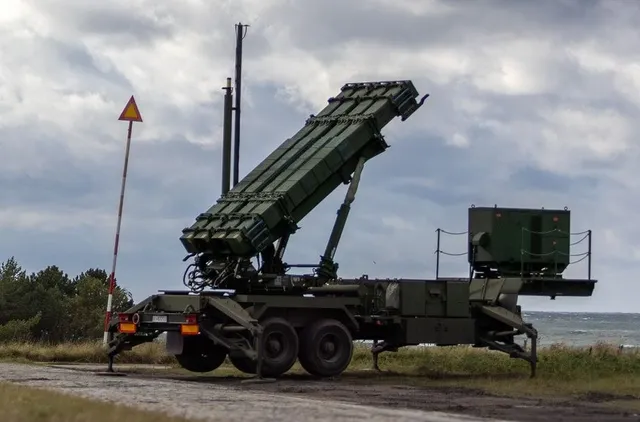 Un vehicul militar verde cu un lansator de rachete înclinat este poziționat pe un drum de pământ sub un cer înnorat.
