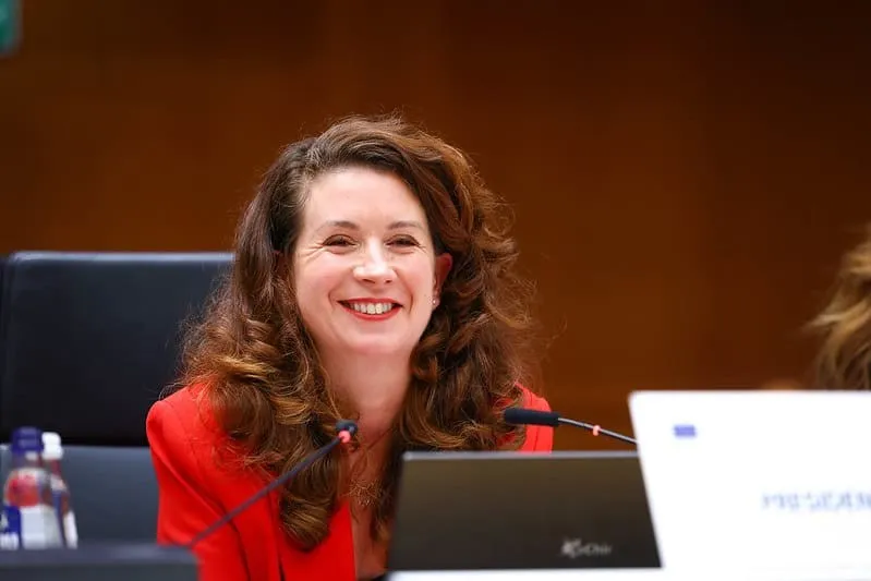 O femeie cu părul castaniu și ondulat poartă un sacou roșu și zâmbește în timp ce stă la o masă cu microfoane și un laptop.