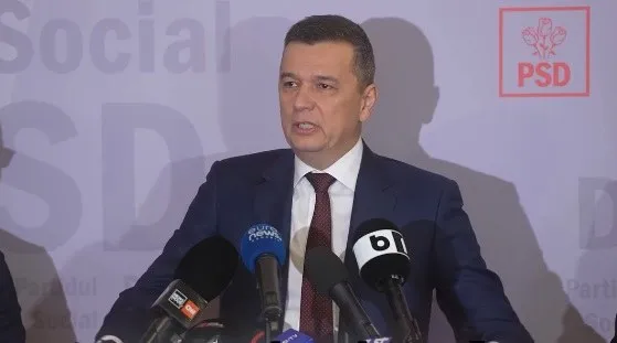 Un bărbat într-un costum închis la culoare și cravată roșie vorbește în fața mai multor microfoane de presă.