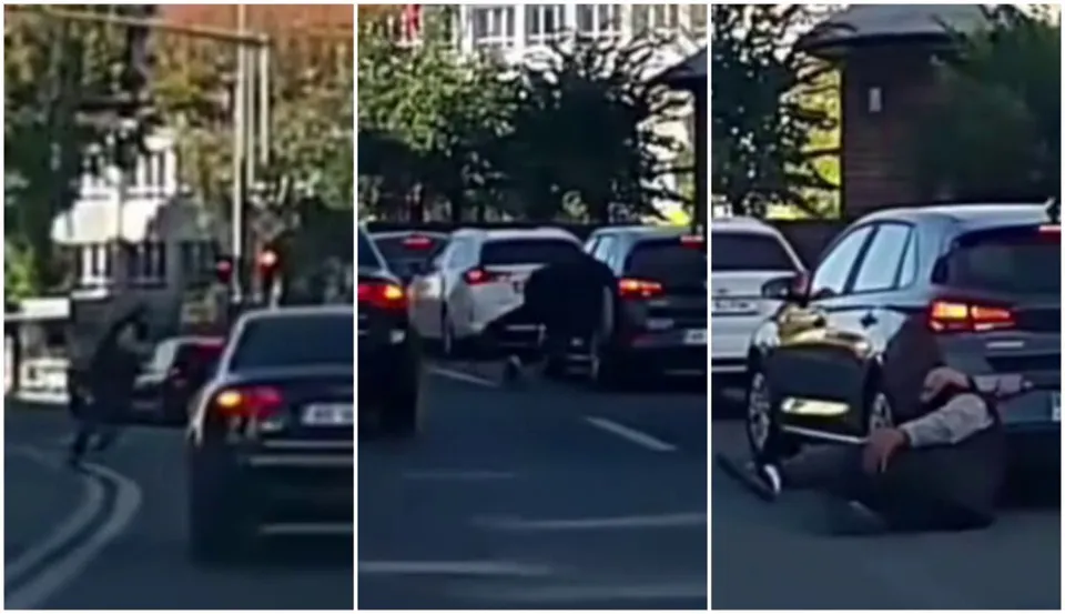 Un bărbat este așezat pe asfalt lângă un autoturism negru pe un drum urban cu alte mașini parcate în fundal.