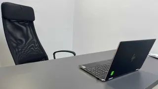 Un laptop deschis și un scaun de birou negru sunt plasate pe o masă gri în fața unui perete alb.