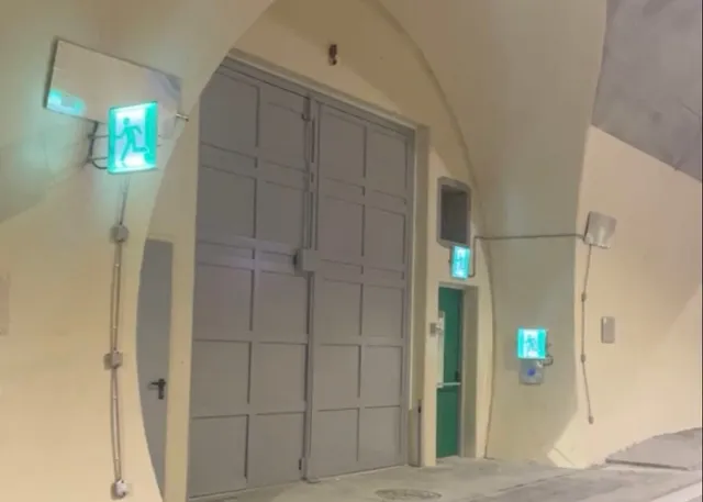 O poartă mare gri este montată într-un perete curbat, lângă ușa verde și semne de ieșire de urgență luminoase pe un fundal de culoare deschisă.