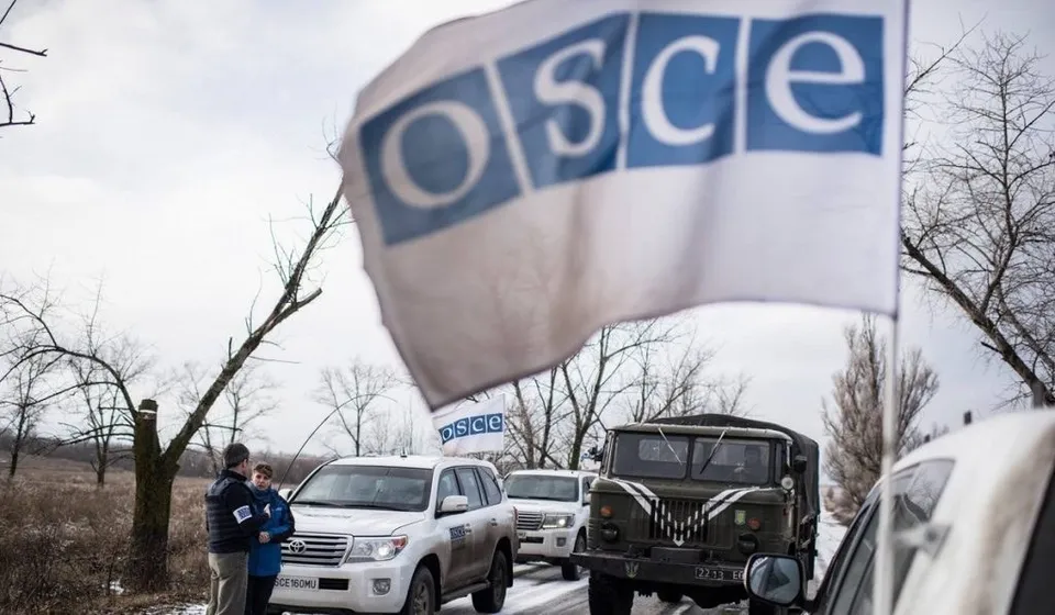 Un steag alb cu logo-ul albastru OSCE flutură în prim-plan, în fața unui grup de mașini parcate pe un drum de țară.