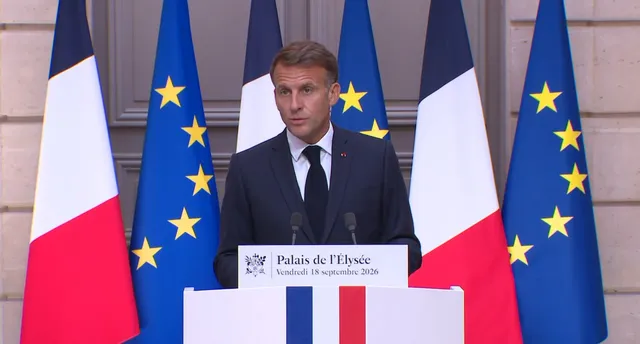 Un bărbat într-un costum închis și cravată neagră stă în spatele unui pupitru alb în fața mai multor steaguri franceze și europene.