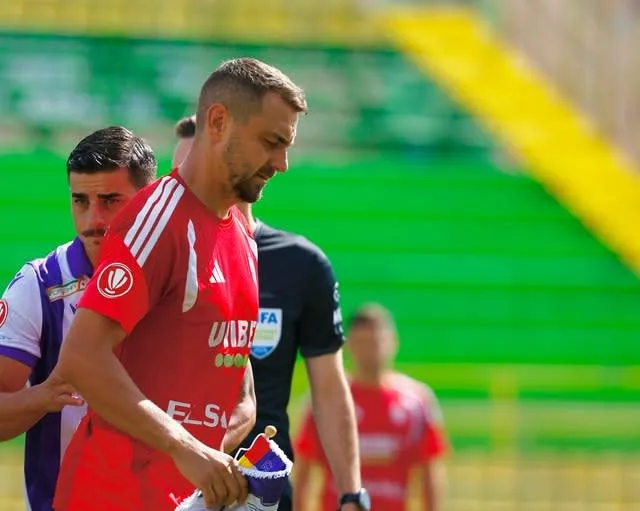 Un jucător de fotbal îmbrăcat în echipament roșu merge pe un teren, fiind însoțit de alți sportivi și un arbitru în fundal.