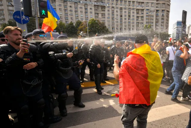Polițiști în echipament de intervenție pulverizează cu gaze lacrimogene spre un protestatar cu steagul României pe umeri, în timpul unei manifestații în stradă.