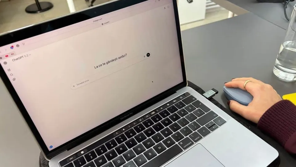 O mână cu un inel subțire ține un mouse albastru lângă un laptop deschis care afișează o interfață de chat pe un ecran alb.