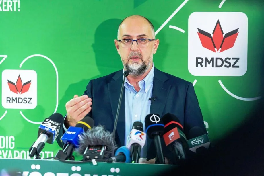 Un bărbat cu ochelari și barbă scurtă poartă un sacou închis și o cămașă deschisă, vorbind în fața mai multor microfoane pe un fundal verde.
