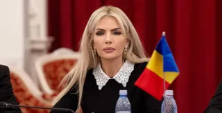 O femeie cu părul blond, purtând o bluză neagră cu guler dantelă albă, stă în fața unui steag tricolor.