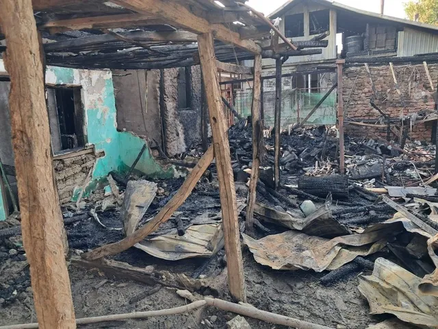 Structura de lemn carbonizată și resturi de metal sunt împrăștiate pe un teren cu cenușă și deșeuri în fața unei clădiri avariate.