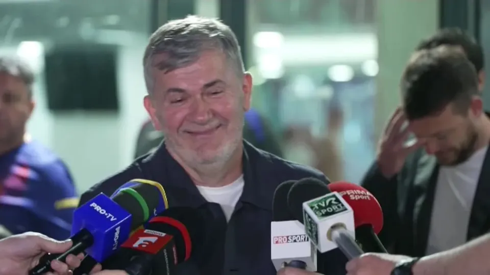 Un bărbat cu părul scurt și gri zâmbește în timp ce mai mulți microfoane de jurnalist sunt îndreptate spre el.