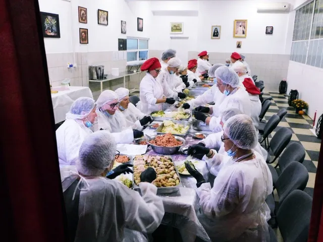 Oameni îmbrăcați în haine albe și bonete de protecție stau la o masă lungă și pregătesc diverse preparate alimentare în containere metalice.