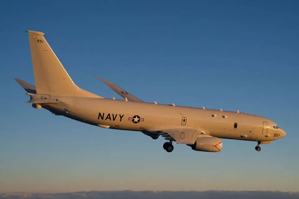 Un avion gri cu inscripția NAVY zboară pe un cer senin deasupra unui orizont îndepărtat.