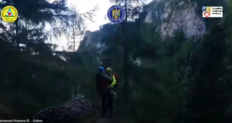 Două persoane purtând echipament de protecție și casci sunt situate într-o zonă împădurită lângă o stâncă sub un cer înnorat.