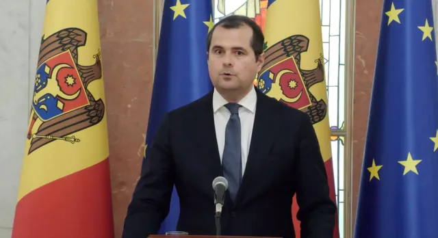 Un bărbat cu cămașă albă, cravată și sacou închis la culoare stă în fața unui microfon, având în spate steaguri colorate.