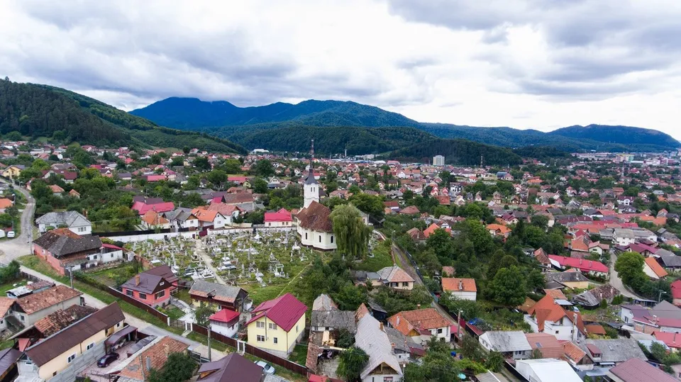 O vedere aeriană a unui oraș cu case cu acoperișuri colorate, copaci verzi și dealuri împădurite sub un cer înnorat.