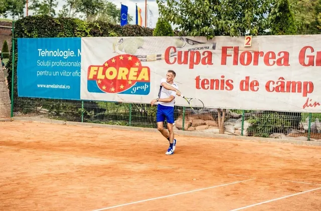 Un bărbat cu tricou alb și pantaloni scurți albaștri lovește o minge cu o rachetă de tenis pe un teren de culoarea cărămizii.