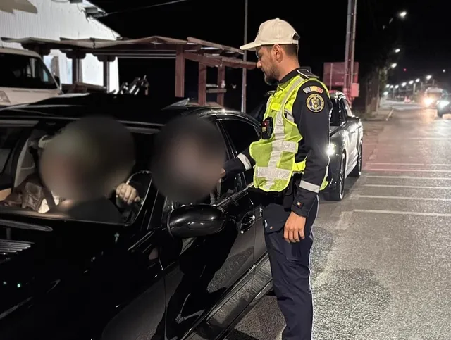 Un polițist în uniformă și vestă reflectorizantă stă lângă portiera unui autoturism negru pe un drum asfaltat noaptea.