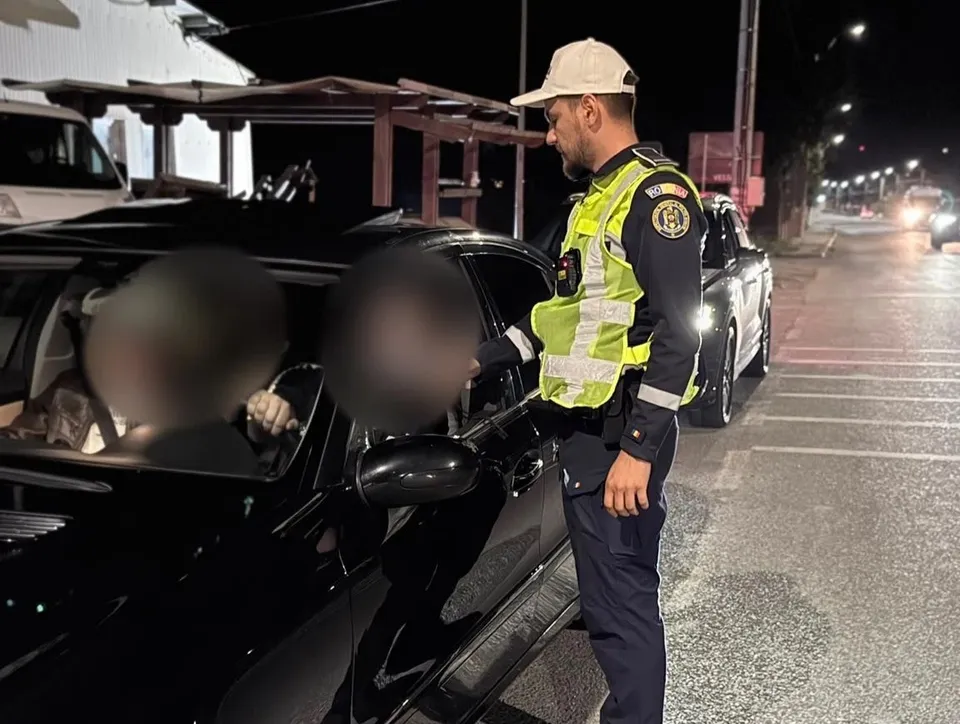Un polițist în uniformă și vestă reflectorizantă stă lângă portiera unui autoturism negru pe un drum asfaltat noaptea.