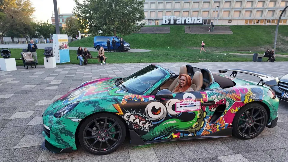 O mașină sport colorată cu grafică este parcată pe o suprafață pavată, având o femeie așezată la volan.