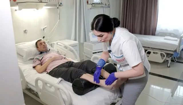 O femeie cu părul negru și cămașă albă aplică un dispozitiv pe picioarele unui bărbat care stă întins pe un pat spitalicesc.