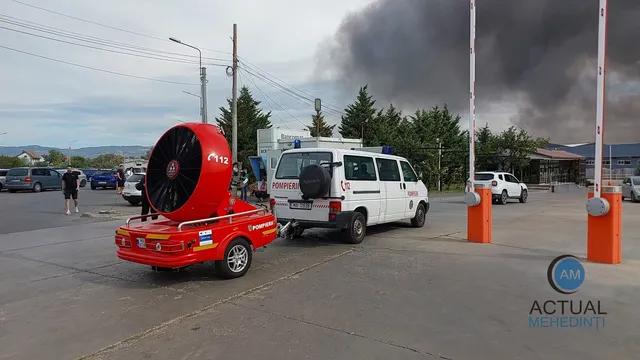 O autospecială roșie cu un ventilator mare este parcată pe un teren asfaltat lângă un van alb și două bariere de acces albe și portocalii.