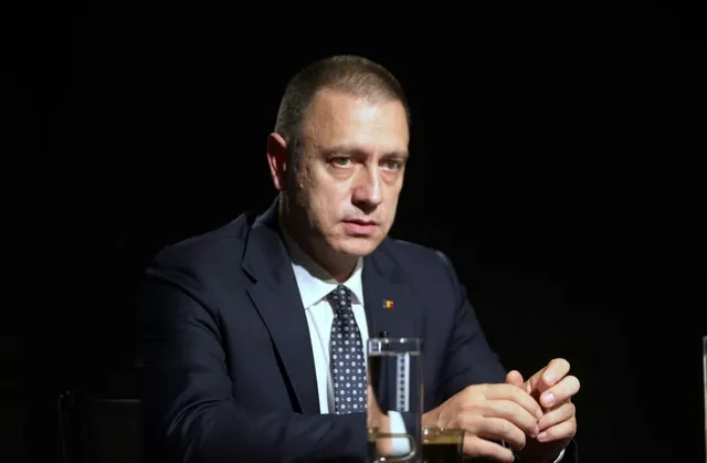 Un bărbat cu părul scurt, îmbrăcat în costum închis și cămașă albă, stă la o masă cu un pahar de sticlă în față.