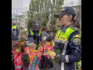 O polițistă în uniformă neagră și galbenă stă lângă un grup de copii mici care poartă tricouri colorate în exterior.