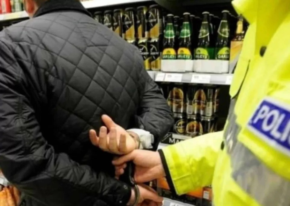 Un agent de poliție în uniformă galbenă aplică cătușe unui bărbat care poartă o jachetă neagră matlasată în fața unor rafturi cu sticle de băuturi.