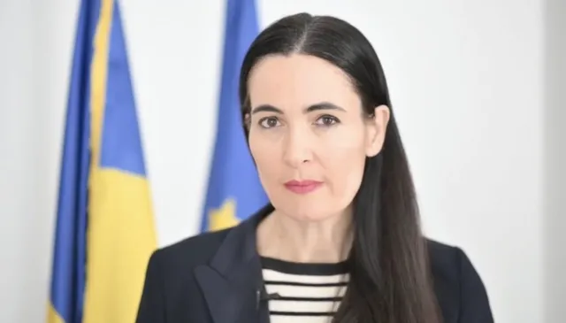 O femeie cu părul lung și negru poartă o jachetă închisă la culoare peste un tricou cu dungi orizontale negre și albe, în fața unor steaguri.