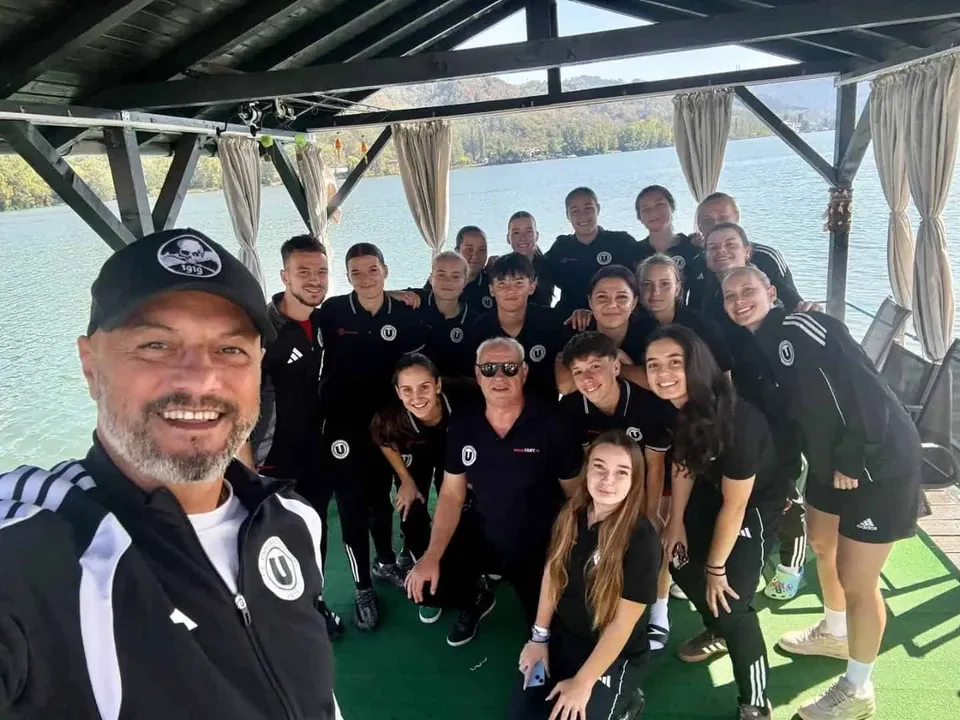 Un grup de persoane îmbrăcate în echipamente sportive negre posedă pentru o fotografie sub o terasă acoperită, lângă un lac.