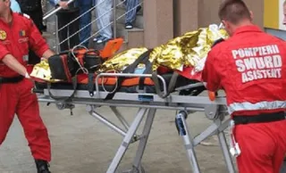 Două persoane în uniforme roșii transportă un cadru medical cu o persoană acoperită de o folie reflectorizantă pe o targă metalică.