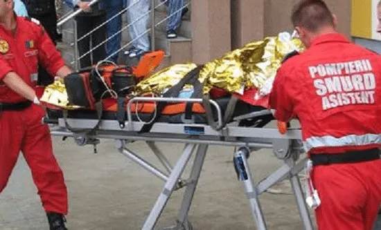 Două persoane în uniforme roșii transportă un cadru medical cu o persoană acoperită de o folie reflectorizantă pe o targă metalică.