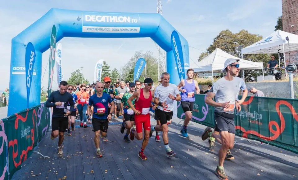 Oamenii în echipament sportiv aleargă pe o pistă de lemn sub o poartă gonflabilă albastră sub cerul senin.