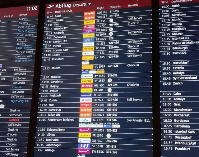 Un ecran digital afișează o listă de zboruri de plecare cu destinații, numere de zbor și orele de decolare pe un fundal negru.
