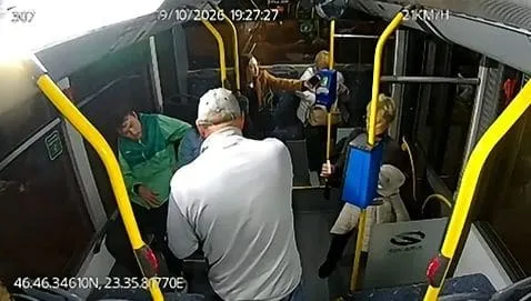 Oameni stau în interiorul unui vehicul lângă barele de prindere galbene, sub o lumină albă intensă.
