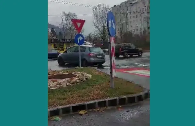 Un autoturism gri este oprit pe o porțiune de drum lângă indicatoare rutiere și un rond de pietre.